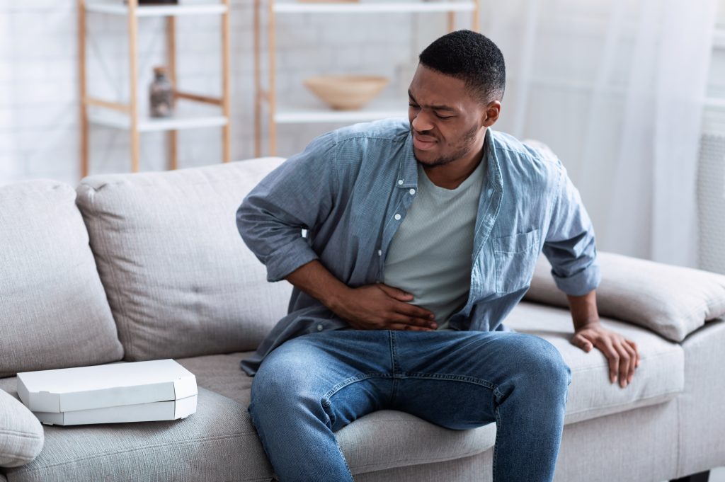 Consumer Health: Recognizing the signs of irritable bowel syndrome a young Black man sitting on a couch, near a pizza box, grimacing in pain, with his hand on his belly
