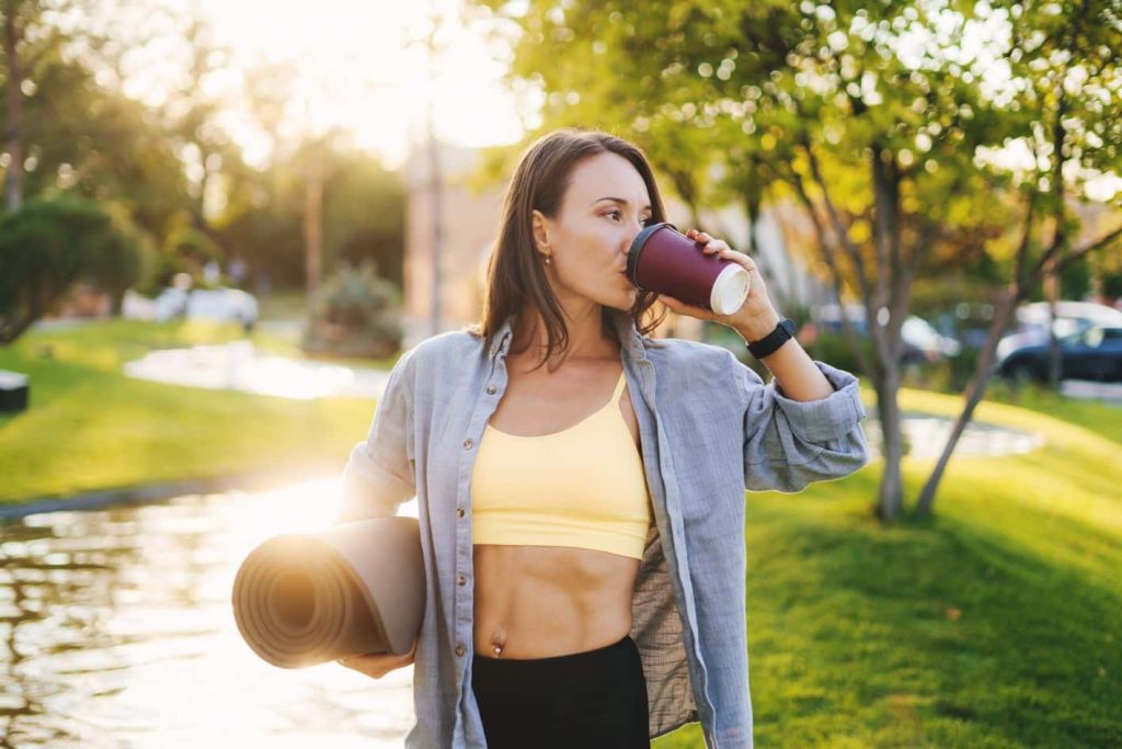 Understanding the Effects of Caffeine Before Workout A woman wearing a yellow sports bra and open button-down shirt is standing outdoors with a yoga mat under her arm, drinking from a disposable cup. She is in a park-like setting with green grass, trees, and a pond in the background. MyFitnessPal Blog