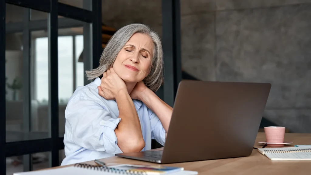 Woman holding her neck at a laptop, showing chronic pain interrupting daily work